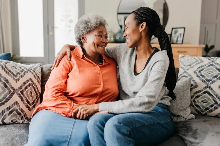 Young woman hugging mother on sofa at home, smiling cheerfully at each other